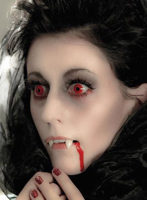 Eine Person mit blasser Haut, roten Augen, die durch die Jahreslinsen „Red Devil Rot“ noch stärker hervorgehoben werden, Vampirzähnen und Kunstblut auf den Lippen; sie trägt ein schwarzes Kostüm und dunkles Make-up und blickt zur Seite.