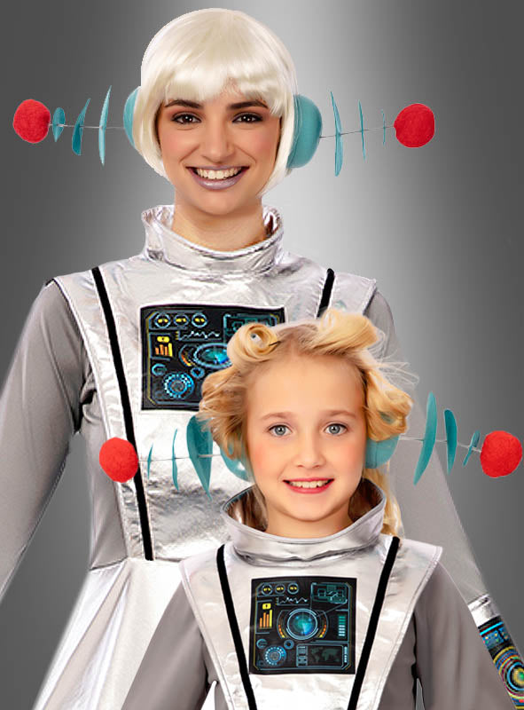 Zwei Personen in silbernen Kostümen im Weltraum-Stil mit futuristischen Kopfbedeckungen – wie zum Beispiel dem schillernden Haarreif mit Alien-Antennen und Motiven, die an Schaltkreise erinnern – stehen nebeneinander und lächeln vor einem grauen Hintergrund.
