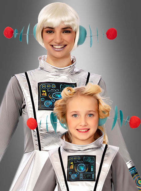 Zwei Personen in silbernen Kostümen im Weltraum-Stil mit futuristischen Kopfbedeckungen – wie zum Beispiel dem schillernden Haarreif mit Alien-Antennen und Motiven, die an Schaltkreise erinnern – stehen nebeneinander und lächeln vor einem grauen Hintergrund.