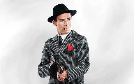 Ein Mann in einem Nadelstreifenanzug und einem schwarzen Hut, der eine Tommy-Gun in der Hand hält, eine Zigarre im Mund, vor einem schlichten Hintergrund stehend.
