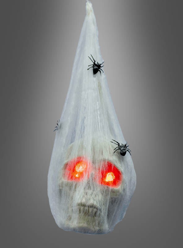 Der „Totenkopf im Spinnenkokon mit LED, 35 cm“ ist in ein weißes Spinnennetz gehüllt und mit Kunststoffspinnen verziert; die leuchtenden Augen sorgen vor dem schlichten grauen Hintergrund für eine besonders gruselige Halloween-Dekoration.