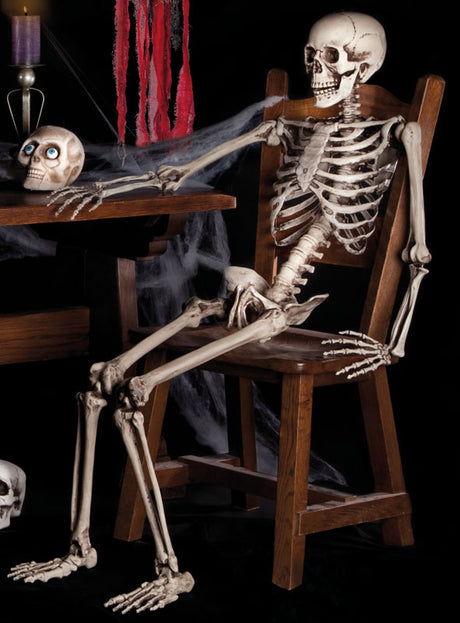 Ein lebensgroßes, 160 cm großes, verstellbares Skelett sitzt auf einem Holzstuhl an einem Tisch, auf dem ein Schädel liegt, dazu Spinnwebendekoration und eine lila Kerze – die perfekte Halloween-Dekoration für eine unvergesslich gruselige Atmosphäre.