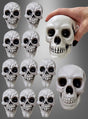 Ein Set aus 12 kleinen, grauen, strukturierten Totenkopf-Halloween-Dekorationsfiguren aus Kunststoff ist in einem Raster angeordnet, wobei oben rechts eine Hand eines der größeren Teile hält – perfekt für Ihre Halloween-Dekoration.