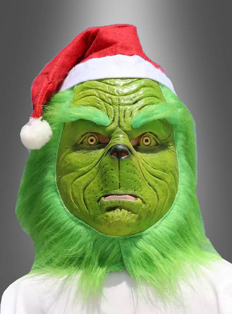 Eine Person, die die „Grünes Weihnachtsmonster“-Vollmaske und eine rote Weihnachtsmannmütze trägt – mit grünem Fell und einem ausdruckslosen Gesicht –, präsentiert vor einem grauen Hintergrund mit Farbverlauf das perfekte Weihnachtskostüm.