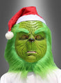 Eine Person, die die „Grünes Weihnachtsmonster“-Vollmaske und eine rote Weihnachtsmannmütze trägt – mit grünem Fell und einem ausdruckslosen Gesicht –, präsentiert vor einem grauen Hintergrund mit Farbverlauf das perfekte Weihnachtskostüm.