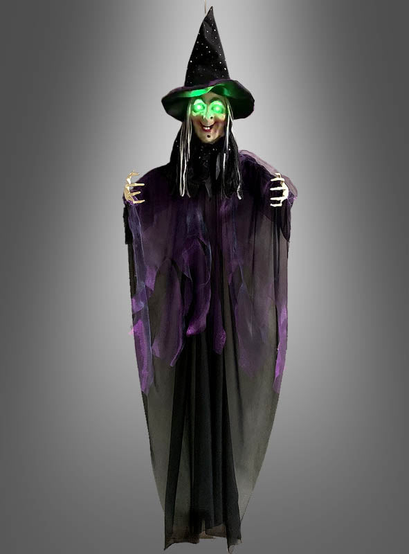 Eine Halloween-Dekoration mit der „Sprechenden Hexe“ (183 cm) mit LED-Augen: Diese Hexe steht vor einem grauen Hintergrund und hat leuchtend grüne LED-Augen, einen schwarzen Spitzhut und eine lange schwarz-lila Robe.