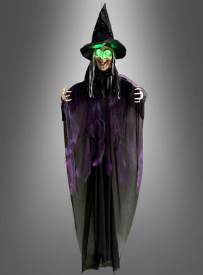 Eine Halloween-Dekoration mit der „Sprechenden Hexe“ (183 cm) mit LED-Augen: Diese Hexe steht vor einem grauen Hintergrund und hat leuchtend grüne LED-Augen, einen schwarzen Spitzhut und eine lange schwarz-lila Robe.