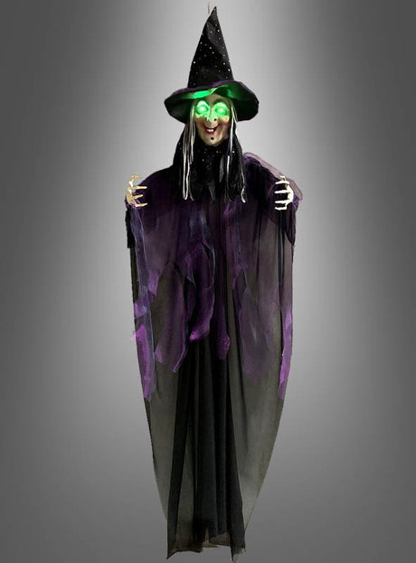 Eine Halloween-Dekoration mit der „Sprechenden Hexe“ (183 cm) mit LED-Augen: Diese Hexe steht vor einem grauen Hintergrund und hat leuchtend grüne LED-Augen, einen schwarzen Spitzhut und eine lange schwarz-lila Robe.