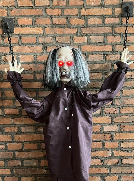 Wir präsentieren den „Lebensgroßen Zombie 185 cm an Ketten animiert“ – eine furchterregende Halloween-Dekoration mit leuchtend roten Augen, verfilztem grauen Haar und Ketten an den Handgelenken, die an einer Backsteinwand befestigt ist.