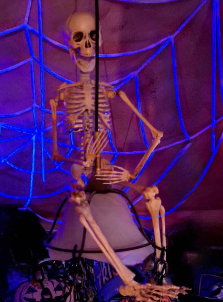 Eine Skelett-Dekoration aus Kunststoff thront auf einer Leuchte, vor dem Hintergrund des „Riesigen UV-Spinnennetzes 300 cm, weiß“ – so entsteht eine gruselige Halloween-Dekoration, die durch violette Beleuchtung und weiße Spinnennetz-Details noch verstärkt wird.