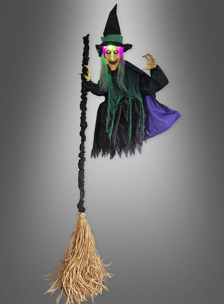 Ein Highlight der Halloween-Dekoration: Die fliegende Hexe mit Besen (210 cm) – mit leuchtend grünen Augen, langen grünen Haaren, einem klassischen schwarzen Hut und einem dramatischen lila Umhang präsentiert sie sich mit ihrem Hexenbesen vor einem stimmungsvollen grauen Hintergrund.