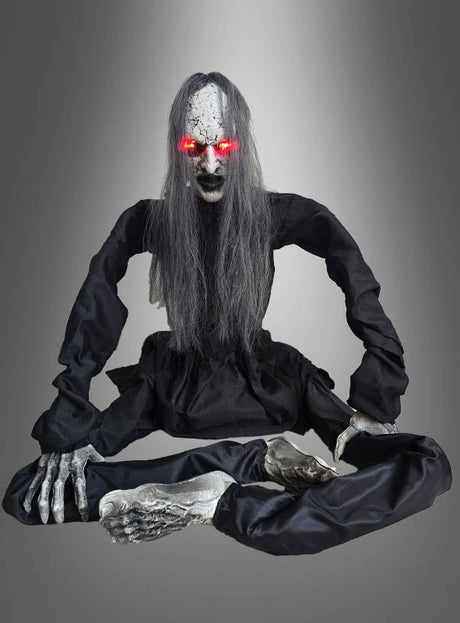 Eine lebensechte Halloween-Requisite in Skelettform mit langen grauen Haaren, leuchtend roten Augen, einem schwarzen Kleid und langgestreckten Gliedmaßen, die mit gekreuzten Beinen vor einem grauen Hintergrund mit Farbverlauf sitzt – diese „Zombie-Figur 60 cm animiert“ wird Ihnen garantiert einen Schauer über den Rücken jagen.