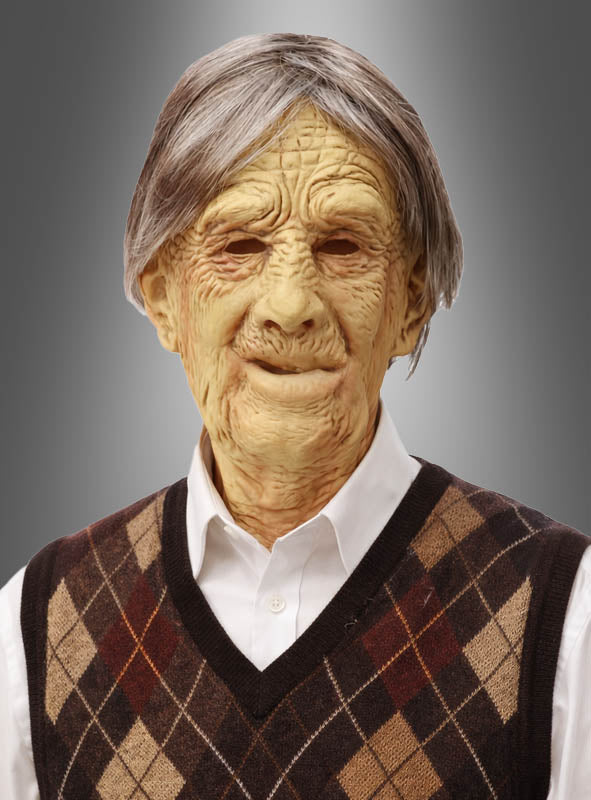 Ein älterer Mann, der eine Strickweste mit Argyle-Muster und ein weißes Hemd trägt, blickt mit neutralem Gesichtsausdruck nach vorne vor einem grauen Farbverlaufshintergrund. Der realistische Look wird durch die Verwendung der „Alter Mann Opa Latex-Maske“ erzielt, die für ein authentisches „Alter Mann“-Erscheinungsbild sorgt.