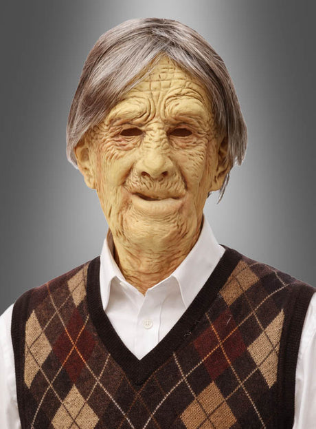 Ein älterer Mann, der eine Strickweste mit Argyle-Muster und ein weißes Hemd trägt, blickt mit neutralem Gesichtsausdruck nach vorne vor einem grauen Farbverlaufshintergrund. Der realistische Look wird durch die Verwendung der „Alter Mann Opa Latex-Maske“ erzielt, die für ein authentisches „Alter Mann“-Erscheinungsbild sorgt.