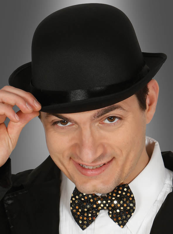 Ein Mann mit einer schwarzen „Gentleman Melone“ und einer mit Pailletten besetzten Fliege lächelt, während er vor einem grauen Hintergrund seinen Hut lüftet.