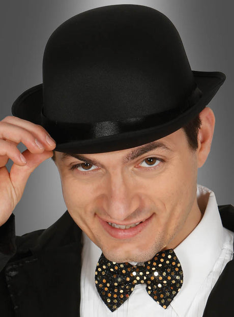 Ein Mann mit einer schwarzen „Gentleman Melone“ und einer mit Pailletten besetzten Fliege lächelt, während er vor einem grauen Hintergrund seinen Hut lüftet.