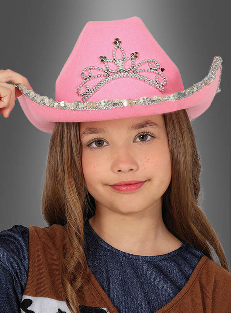 Ein junges Mädchen, das einen rosa Cowboyhut für Kinder mit einem silbernen Tiara-Motiv und funkelnden Pailletten trägt, blickt mit ausdruckslosem Gesicht in die Kamera.