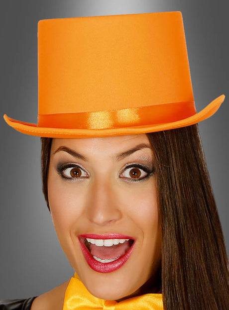 Eine Frau, die den „Orangener Zylinder Hut Unisex“ und eine dazu passende Fliege trägt, mit großen Augen und offenem Mund, steht vor einem grauen Hintergrund – perfekt für jeden Anlass, bei dem ein Showkostüm gefragt ist.
