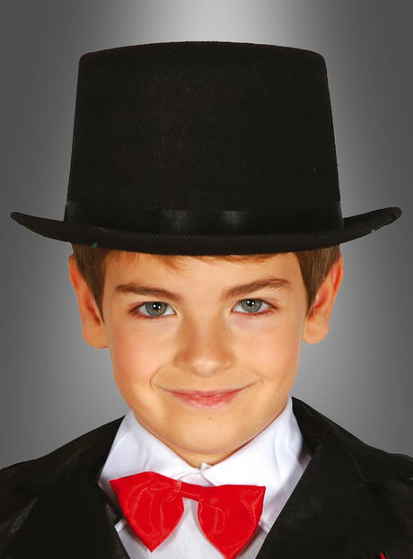 Ein kleiner Junge, der den „Schwarzen Zylinder“ für Kinder trägt, dazu eine rote Fliege und eine schwarze Jacke – ideal für ein Schornsteinfeger-Kostüm –, lächelt vor einem grauen Hintergrund mit Farbverlauf in die Kamera.