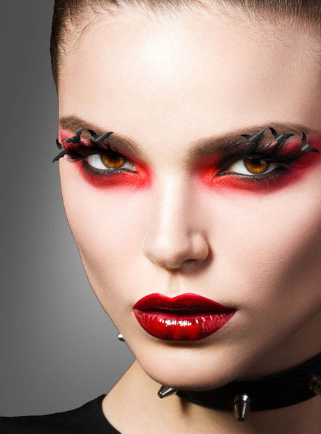 Eine Frau mit auffälligem rotem Augen-Make-up, glänzendem rotem Lippenstift und aufwendigen „Fledermaus Wimpern für Vamps“ – künstliche Wimpern in Form schwarzer Vögel –, kombiniert mit einem schwarzen Stachel-Halsband, blickt vor einem grauen Hintergrund nach vorne. Abgerundet wird der Look durch diese gotischen „Fledermaus-Wimpern für Vamps“, die für einen zusätzlichen Hauch dunkler Eleganz sorgen.