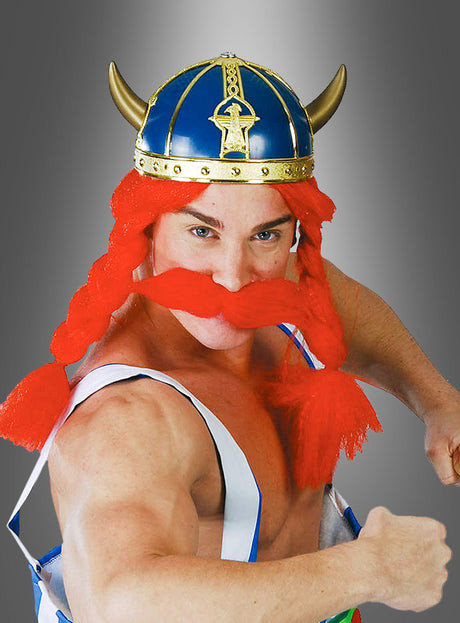 Eine kostümierte Person mit einem blauen Wikingerhelm für Männer, einer rot geflochtenen Perücke und einem roten Kunstschnurrbart, die Hosenträger trägt und mit erhobenen Fäusten vor einem grauen Hintergrund posiert.