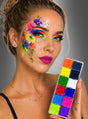 Eine Frau mit bunter Blumen-Gesichtsbemalung hält eine bunte Make-up-Palette mit 12 Farben in der Hand und posiert vor einem grauen Hintergrund mit Farbverlauf.