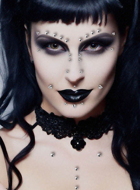 Eine Person mit Gothic-Make-up – schwarzer Lippenstift, auffälliger Lidschatten, ein schwarzes Spitzenhalsband und metallische Gesichtspiercings, die mit den „Fake Piercings zum Aufkleben, 30 Stück“ angebracht wurden – blickt direkt in die Kamera.