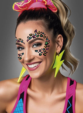 Eine lächelnde blonde Frau trägt neonfarbene Blitz-Ohrringe, ein rosa Haargummi und auffälliges Festival-Make-up, bei dem das „Buntes Leoparden Glitzer-Tattoo fürs Gesicht“ als Gesichtsbemalung dient. Sie trägt bunte Sportkleidung vor einem grauen Hintergrund.
