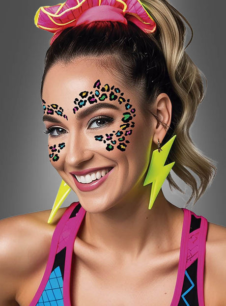 Eine lächelnde blonde Frau trägt neonfarbene Blitz-Ohrringe, ein rosa Haargummi und auffälliges Festival-Make-up, bei dem das „Buntes Leoparden Glitzer-Tattoo fürs Gesicht“ als Gesichtsbemalung dient. Sie trägt bunte Sportkleidung vor einem grauen Hintergrund.