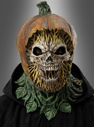 Eine Person trägt die „Gruselige Kürbis-Maske aus Latex“ mit einem Totenkopfgesicht, dunklen Augenlöchern und grünen Blattakzenten, dazu einen schwarzen Umhang vor einem grauen Hintergrund mit Farbverlauf. Diese auffällige Halloween-Maske sorgt für einen unheimlichen und zugleich festlichen Auftritt.