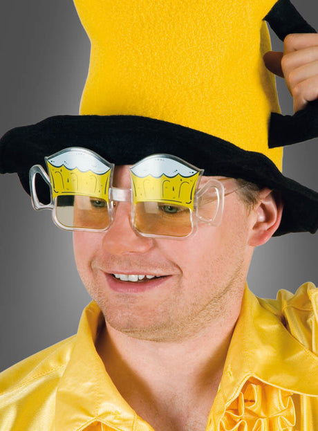 Ein Mann mit einem großen gelben Hut, einem gelben Hemd und einem breiten Lächeln steht vor einem grauen Hintergrund und trägt die „Partybrille Bierkrug Stil Anton“ – eine ausgefallene Brille in Form eines Bierkrugs –, die sich perfekt für jede Oktoberfest-Feier mit Spaßbrillen eignet.