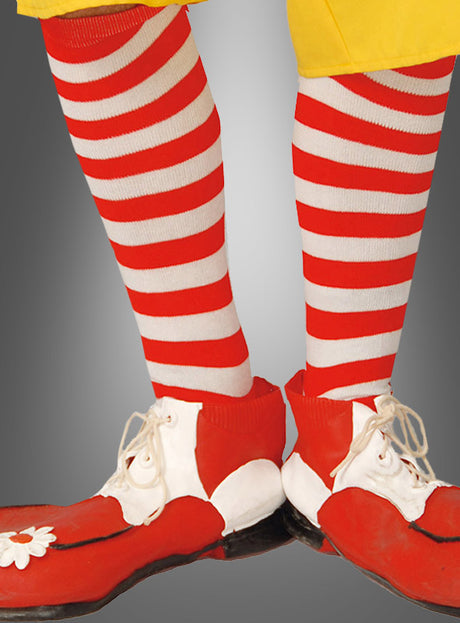 Eine Person, die rot-weiße Ringelstrümpfe und Clownsocken sowie übergroße rot-weiße Clownschuhe zu einer gelben Hose trägt, steht vor einem grauen Hintergrund – ein klassisches Detail jedes Clownskostüms.