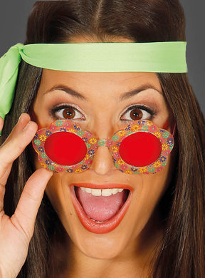 Eine Frau, die die „Runde Blumenbrille für Hippie-Kostüme“ trägt – perfekt als Accessoire für Hippie-Kostüme – mit rotem, blumenförmigem Rahmen und einem grünen Stirnband, schaut überrascht und hat den Mund offen.