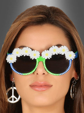 Eine Frau, die eine Brille mit Blumen für Hippies – verziert mit weißen Gänseblümchen – und ein Paar Ohrringe in Form eines Peace-Zeichens trägt, präsentiert klassische Hippie-Accessoires vor einem grauen Farbverlaufshintergrund.