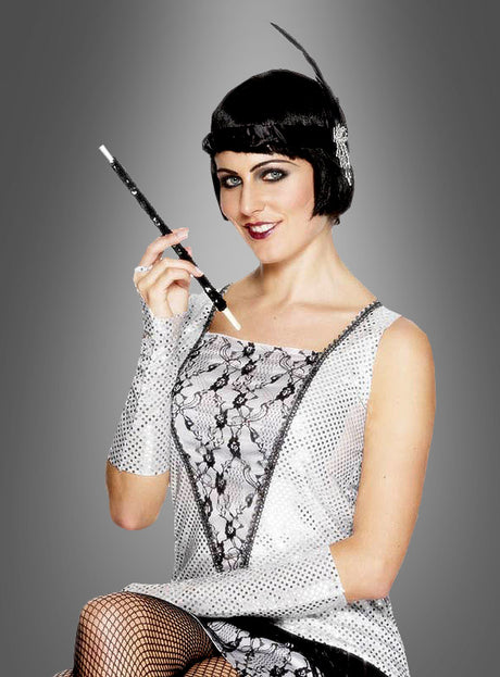 Eine Frau in einem silber-schwarzen Flapperkleid mit passenden Handschuhen und einem Stirnband hält einen Zigarettenhalter aus den 20er Jahren mit Pailletten in der Hand und posiert vor einem grauen Hintergrund.