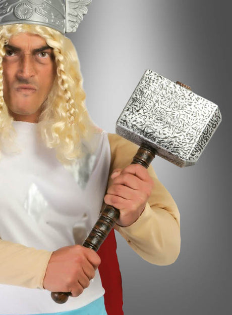Eine Person in einem Kostüm mit einem geflügelten Helm, einer langen blonden Perücke und einem weißen Hemd, die den 50 cm großen „Donnergott“-Superhelden-Hammer vor einem grauen Farbverlaufshintergrund in der Hand hält.