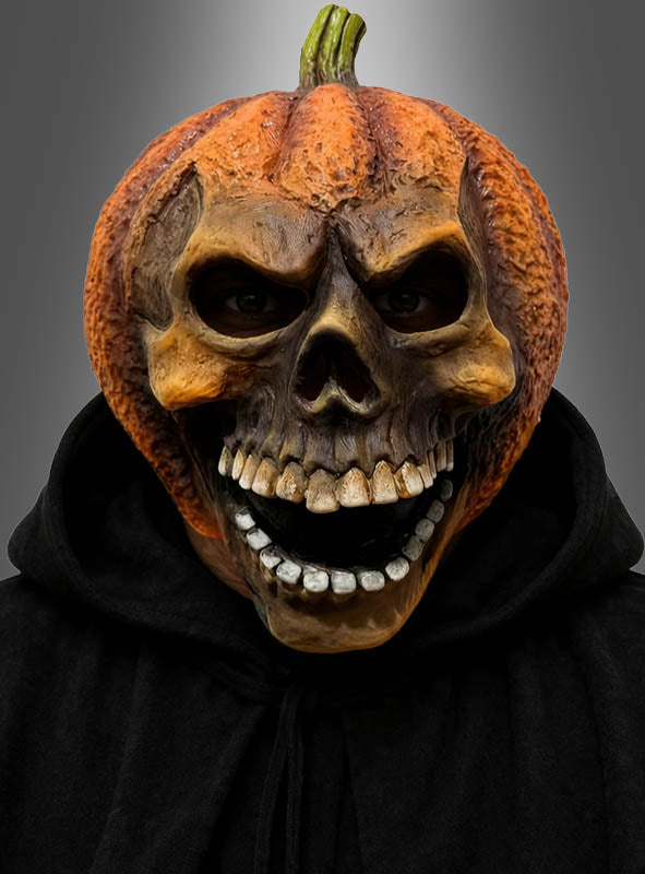 Eine Person, die die „Kürbis-Maske“ – eine lachende Totenkopfmaske aus Latex – und einen schwarzen Kapuzenmantel trägt, blickt vor einem schlichten grauen Hintergrund nach vorne.