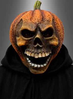 Eine Person, die die „Kürbis-Maske“ – eine lachende Totenkopfmaske aus Latex – und einen schwarzen Kapuzenmantel trägt, blickt vor einem schlichten grauen Hintergrund nach vorne.