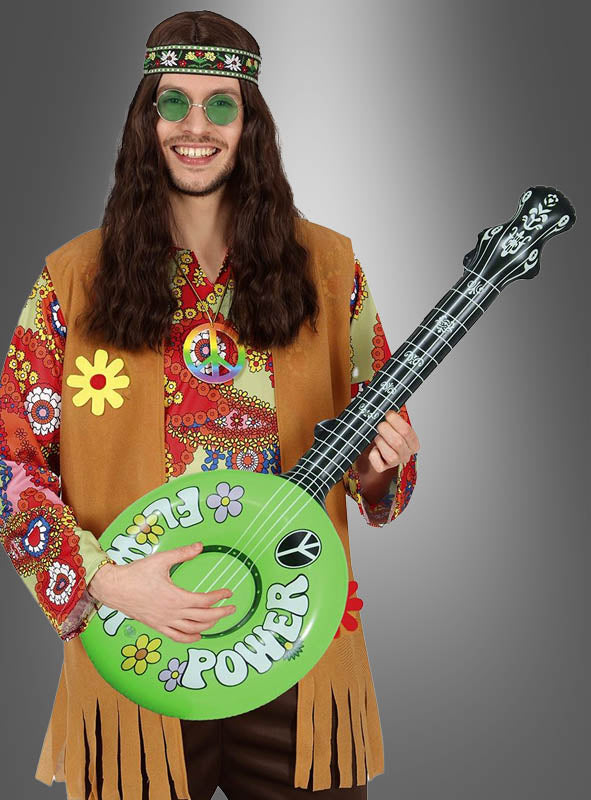 Ein Mann in einem bunten Hippie-Kostüm aus den 1960er Jahren mit langen Haaren und runder Brille, der ein „Aufblasbares Banjo für Hippies“ in der Hand hält, das mit „Flower Power“-Schriftzügen und Friedenssymbolen verziert ist und vor einem grauen Hintergrund steht – perfekt für jede Hippie-Party.
