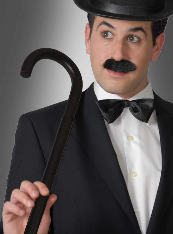 Ein Mann in schwarzem Anzug, mit Fliege und Melone, der einen „Spazierstock schwarz 80 cm“ als perfektes Kostümzubehör in der Hand hält, einen falschen Schnurrbart trägt und vor einem grau verlaufenden Hintergrund steht.