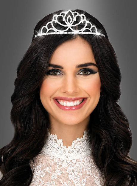 Eine Frau mit langen dunklen Haaren trägt die „Diadem Strass Krone aus Metall“, eine funkelnde Krone, die mit Strasssteinen verziert ist, sowie ein weißes Spitzenkleid. Vor einem grauen Hintergrund lächelt sie in die Kamera.