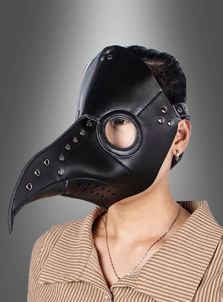 Eine Person, die die „Deluxe Pest Maske“ trägt – gefertigt aus schwarzem Kunstleder mit runden Glasaugenöffnungen und Nietenverzierungen –, steht vor einem grauen Hintergrund mit Farbverlauf.