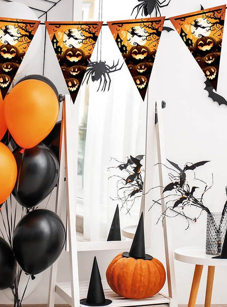 Der Raum ist für Halloween mit orangefarbenen und schwarzen Luftballons, einem Kürbis mit Hexenhut, Wimpelketten mit Halloween-Motiven – darunter die „Lustige Wimpelgirlande 300 cm Halloween-Kürbisse“ – sowie künstlichen Zweigen, die in einer Vase arrangiert sind, dekoriert.