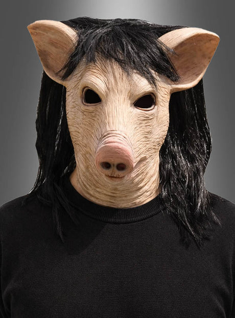 Eine Person, die die „Schwein Latex-Maske mit Haaren“ mit schwarzen Haaren trägt und dazu ein schwarzes Shirt, steht vor einem neutralen grauen Hintergrund. Inspiriert vom gruseligen Aussehen der „SAW“-Maske sorgt dieses unheimliche Halloween-Accessoire für einen wahrhaft gespenstischen Effekt.
