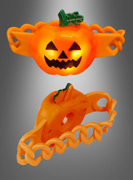 Ein blinkendes Kürbis-Armband aus orangefarbenem Kunststoff mit leuchtendem Jack-O’-Lantern-Gesicht und grünem Stiel, dargestellt in Vorder- und Seitenansicht vor einem grauen Farbverlaufshintergrund.