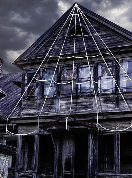 Ein altes, verwittertes Holzhaus mit vernagelten Fenstern ist unter einem bewölkten, dunklen Himmel mit der „Spinnennetz 5 Meter Riesige Halloween-Deko“ – großen weißen Seilen, die ein riesiges Spinnennetz bilden – behängt, was für die perfekte gruselige Atmosphäre sorgt.