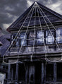 Ein altes, verwittertes Holzhaus mit vernagelten Fenstern ist unter einem bewölkten, dunklen Himmel mit der „Spinnennetz 5 Meter Riesige Halloween-Deko“ – großen weißen Seilen, die ein riesiges Spinnennetz bilden – behängt, was für die perfekte gruselige Atmosphäre sorgt.