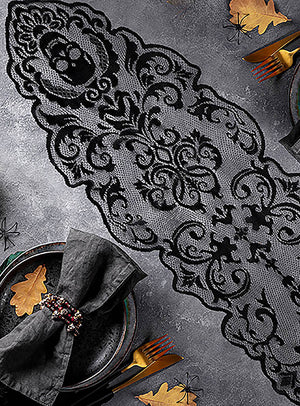 Der Tischläufer „180 x 35 cm – Schwarzer Gothic-Totenkopf“ mit kunstvollen schwarzen Spitzenmustern liegt auf einem grauen Untergrund, umgeben von dunklen Tellern, grauen Servietten, goldenem Besteck und dekorativen Goldblättern – eine elegante Wahl für Ihre Halloween-Tischdekoration.