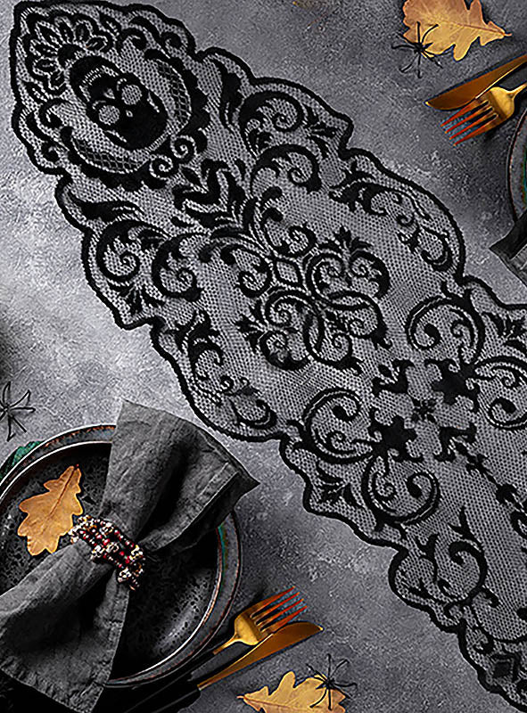 Der Tischläufer „180 x 35 cm – Schwarzer Gothic-Totenkopf“ mit kunstvollen schwarzen Spitzenmustern liegt auf einem grauen Untergrund, umgeben von dunklen Tellern, grauen Servietten, goldenem Besteck und dekorativen Goldblättern – eine elegante Wahl für Ihre Halloween-Tischdekoration.