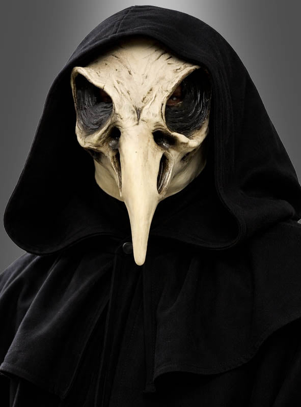 Eine Person, die einen schwarzen Kapuzenmantel und eine „Krähen Schädel“-Halloween-Maske im Stil eines Pestdoktors mit langem Schnabel trägt, steht vor einem schlichten grauen Hintergrund.